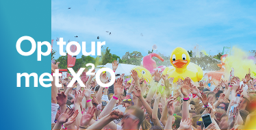 Een uitbundige festivalmenigte onder een blauwe lucht, gooit kleurpoeder in de lucht met een grote opblaasbare badeend in het midden, tijdens de zomertour van X²O badkamers.